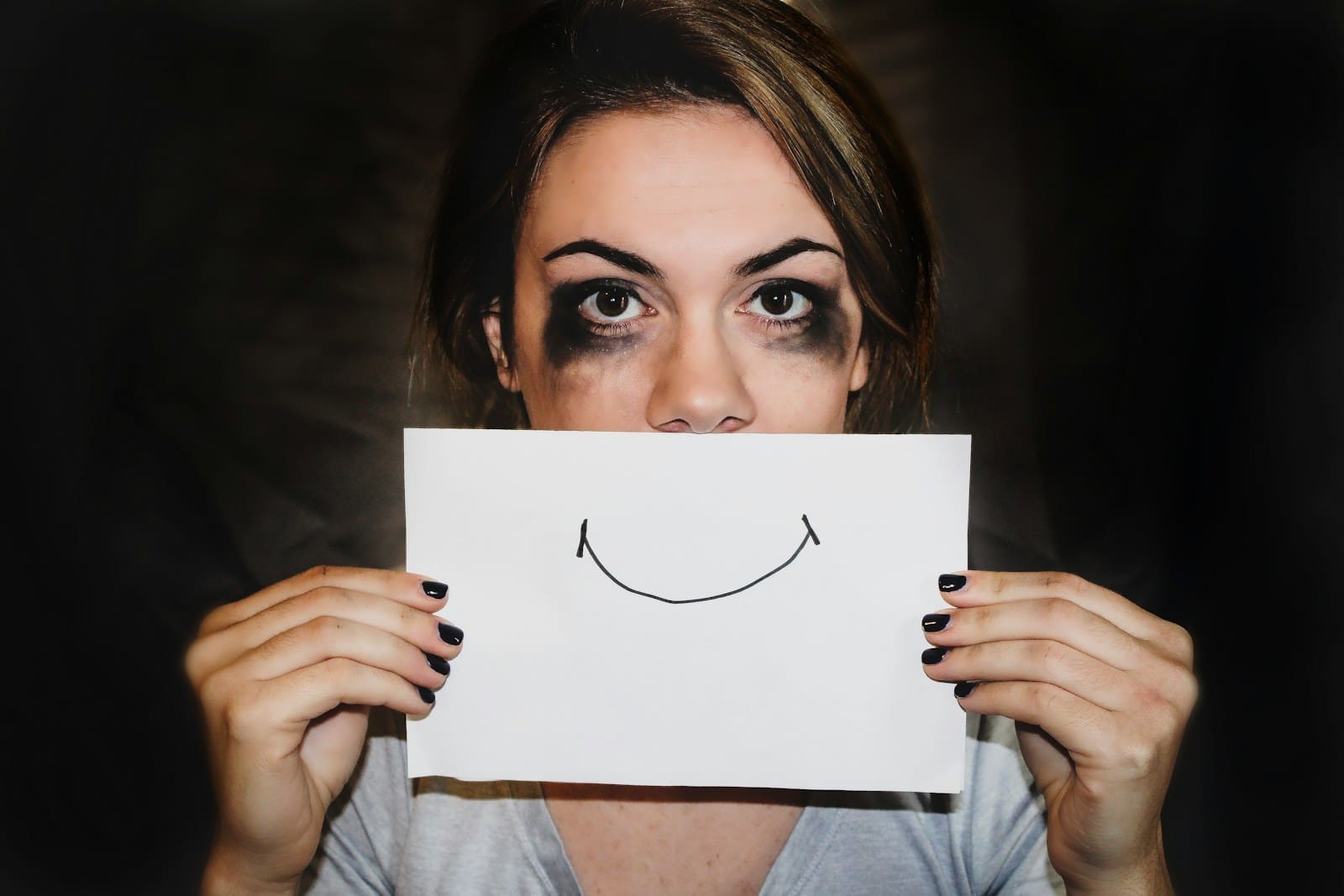person, who appears to have been crying, holding white printer paper with a smile scrawled on it over their mouth